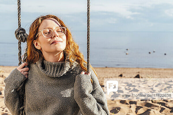 Woman with eyes closed enjoying sunlight on swing at beach