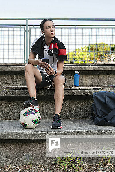 Young woman with soccer ball listening music through in-ear headphones on bench
