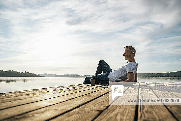 Mature man relaxing by disposable coffee cup on pier by lake