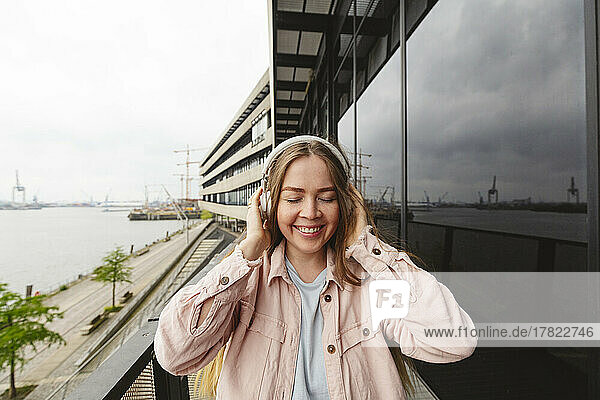 Happy young woman with eyes closed listening music through wireless headphones standing on balcony