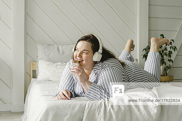 Happy woman eating chocolate and listening music through wireless headphones lying on bed at home