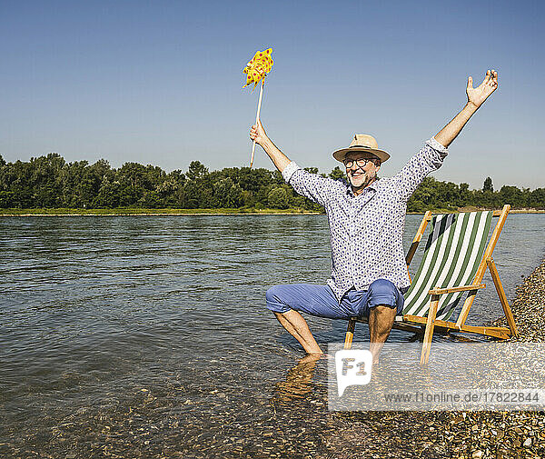 Happy man with arms outstretched holding paper pinwheel toy at riverbank