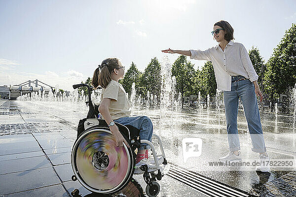 Girl with disability looking at mother playing with splashing fountain water on sunny day