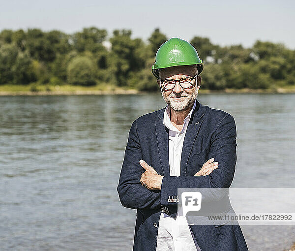 Smiling senior engineer wearing green hardhat standing with arms crossed at riverbank on sunny day