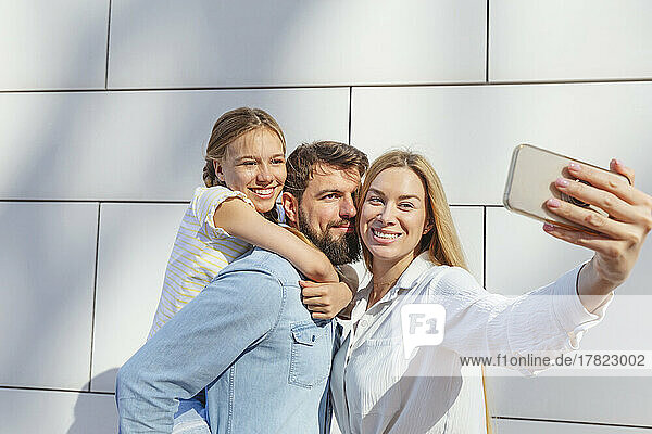 Happy woman taking selfie with family through smart phone in front of wall