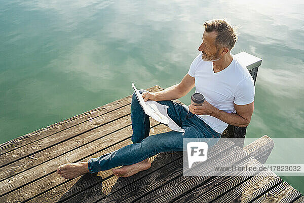 Man with newspaper and disposable coffee cup sitting on pier