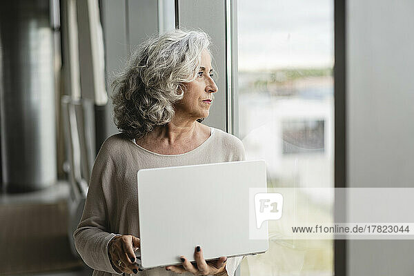 Senior businesswoman with laptop looking out of window at office