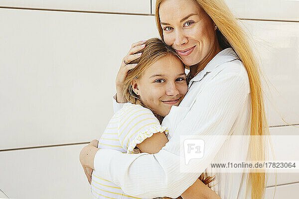 Happy woman with blond hair hugging daughter in front of wall