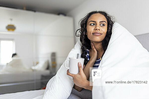 Contemplative woman holding cup of coffee sitting in bed at home