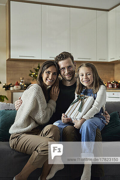 Glückliches Mädchen hält Weihnachtsgeschenk in der Hand und sitzt mit den Eltern zu Hause auf dem Sofa