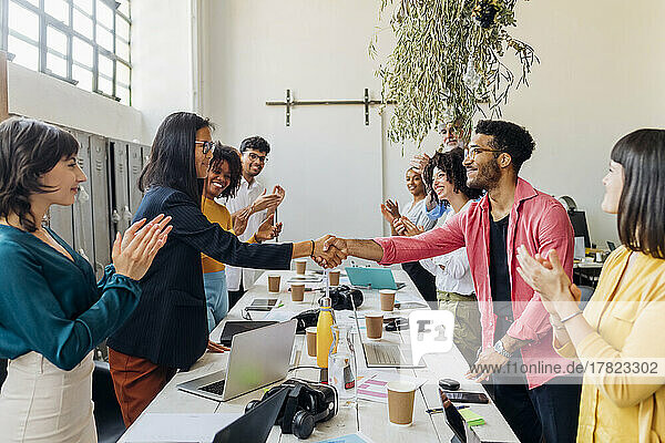 Businessman and businesswoman shaking hands by colleagues at work place