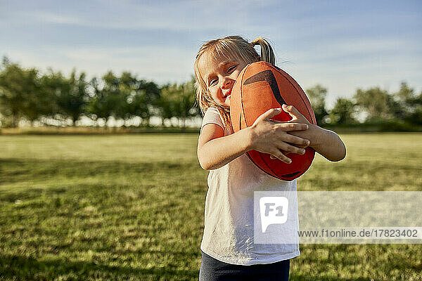 Lächelndes Mädchen umarmt Rugbyball auf dem Sportplatz an einem sonnigen Tag