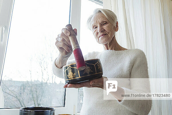 Smiling senior woman with singing bowl standing by window at home
