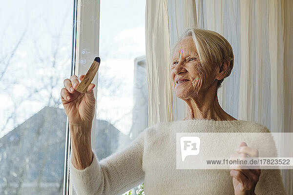 Glückliche ältere Frau mit Palo-Santo-Stick am Fenster
