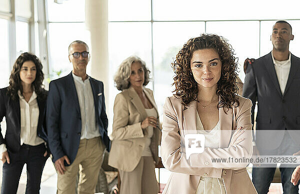 Confident businesswoman with arms crossed standing with colleagues in background at office