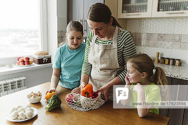 Girls looking at mother taking vegetables from mesh bag in kitchen
