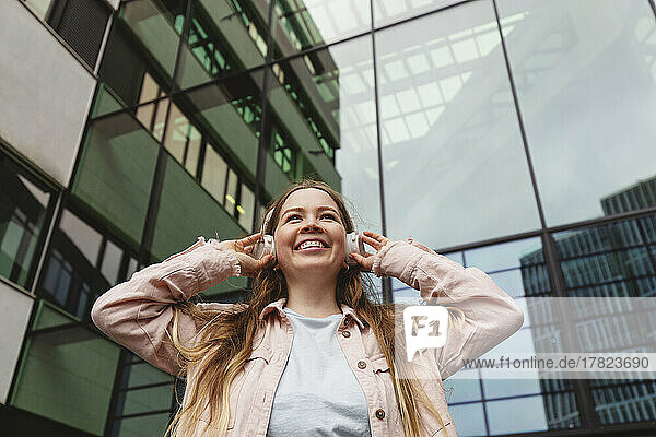 Happy woman wearing wireless headphones standing in front of building