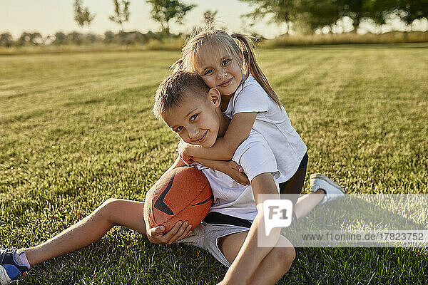 Smiling blond girl hugging brother sitting with rugby ball at sports field