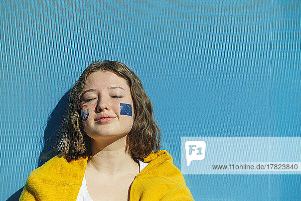 Teenage girl with peace symbol and European Union paint on cheeks in front of blue wall