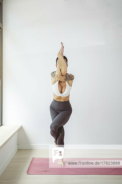 Woman practicing Garudasana on exercise mat in front of wall at yoga studio