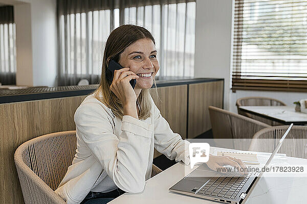 Lächelnde Geschäftsfrau  die mit einem Laptop im Restaurant sitzt und mit dem Handy telefoniert