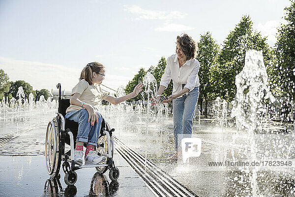 Girl in wheelchair playing along fountain water with mother at park