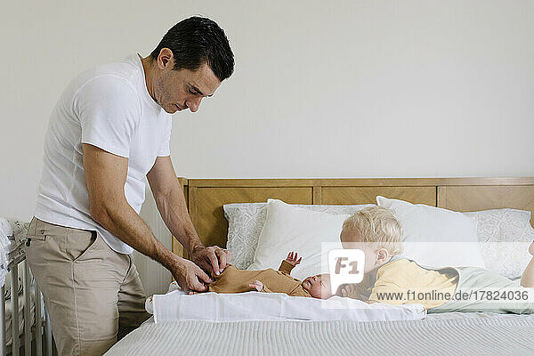 Boy looking at man buttoning baby's rompers on bed at home