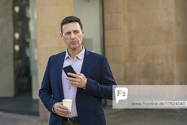 Contemplative businessman holding smart phone and disposable cup