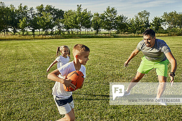 Father playing rugby with son and daughter at sports field on sunny day