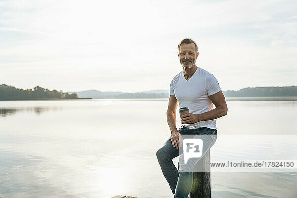Happy mature man with disposable coffee cup sitting on post in front of lake
