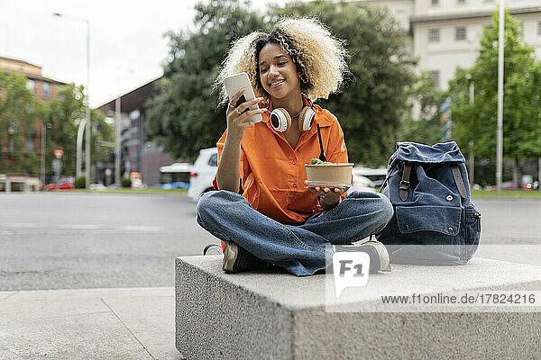 Smiling woman using smart phone holding lunch box sitting cross-legged on seat