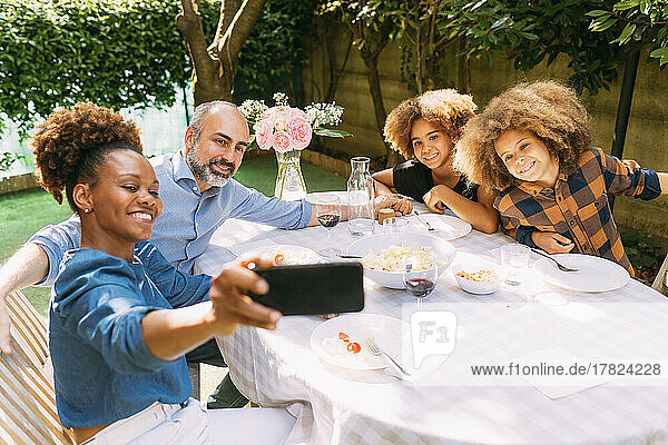 Lächelnde Frau macht ein Selfie mit der Familie per Smartphone am Esstisch im Hinterhof