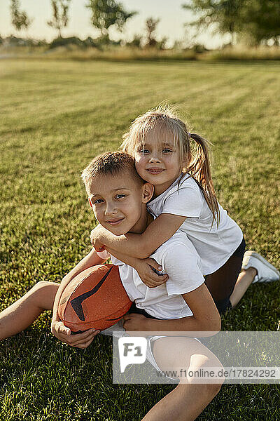 Smiling girl hugging brother sitting with rugby ball at sports field on sunny day