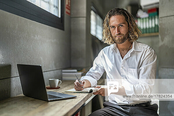Serious businessman with pen and diary by laptop at table in office