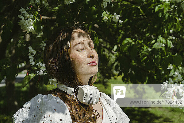 Woman enjoying sunlight with headphones around neck in park