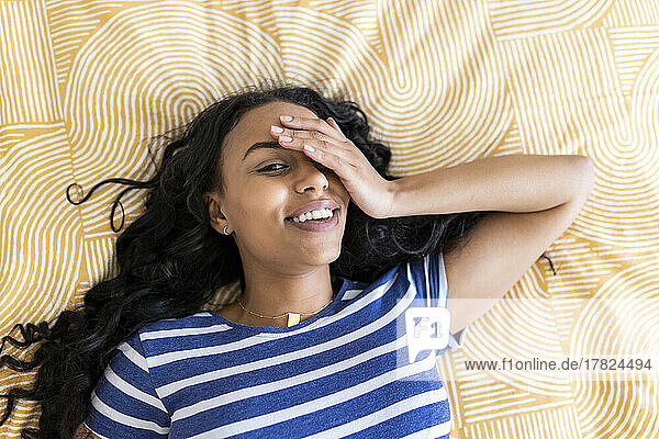 Smiling woman covering eye with hand lying on bed at home