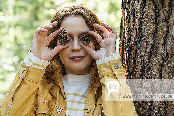 Smiling woman covering eyes with pine cone in forest