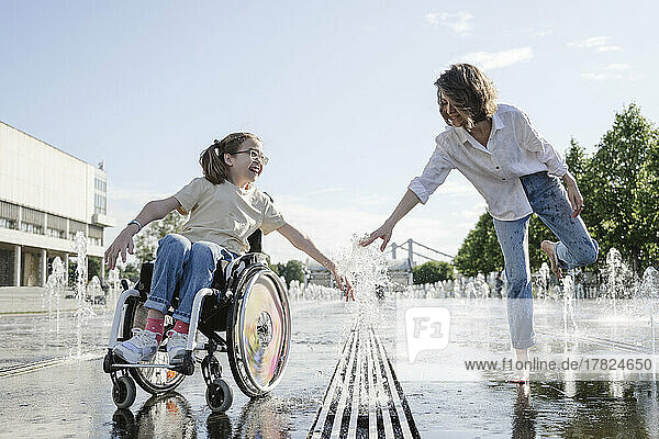 Woman and daughter with disability playing along fountain at park on sunny day