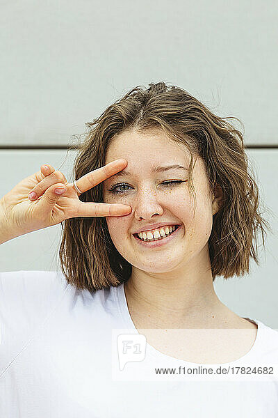 Fröhliches Teenager-Mädchen zwinkert mit dem Auge und gestikuliert ein Friedenszeichen vor der Wand