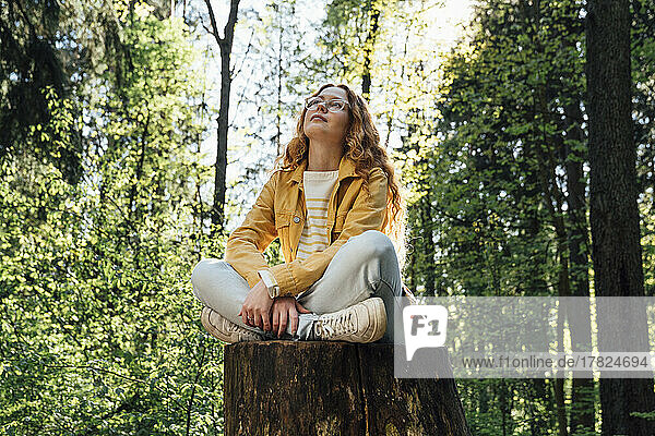 Thoughtful woman sitting cross-legged on tree stump in forest