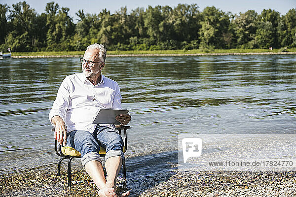 Senior man holding tablet PC sitting on chair at riverbank