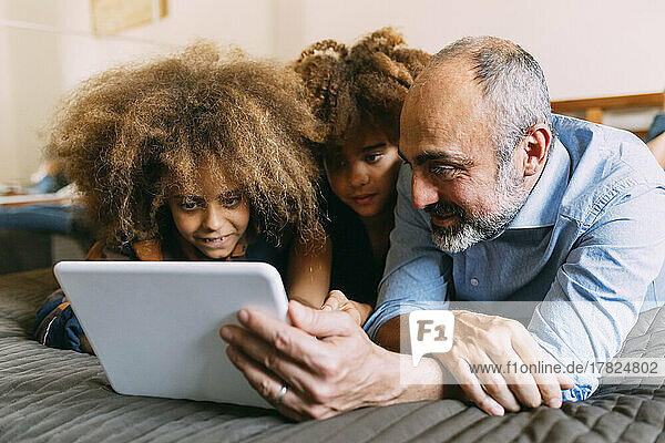 Smiling man sharing tablet PC with children lying on bed at home