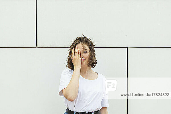 Teenage girl covering eye standing in front of wall