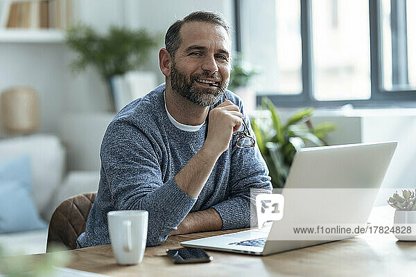 Smiling freelancer holding eyeglasses sitting with laptop at home