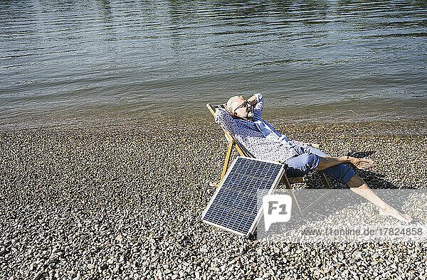 Senior man with hands behind head sitting by solar panel at riverbank