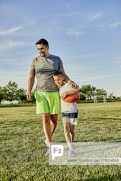 Smiling father and son walking together at sports field on sunny day