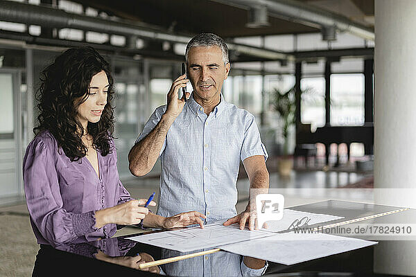 Businessman talking on smart phone standing with colleague looking at documents in office