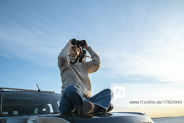 Smiling young woman looking through binoculars sitting on car hood