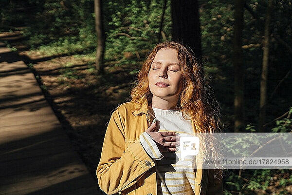 Young woman with hand on chest enjoying sunlight in forest