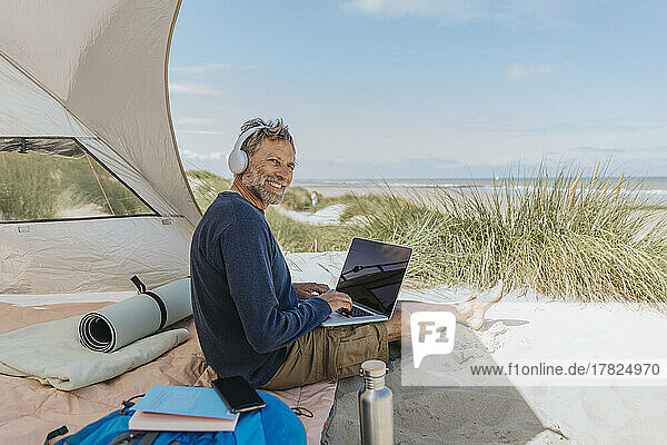 Happy freelancer wearing wireless headphones sitting at beach